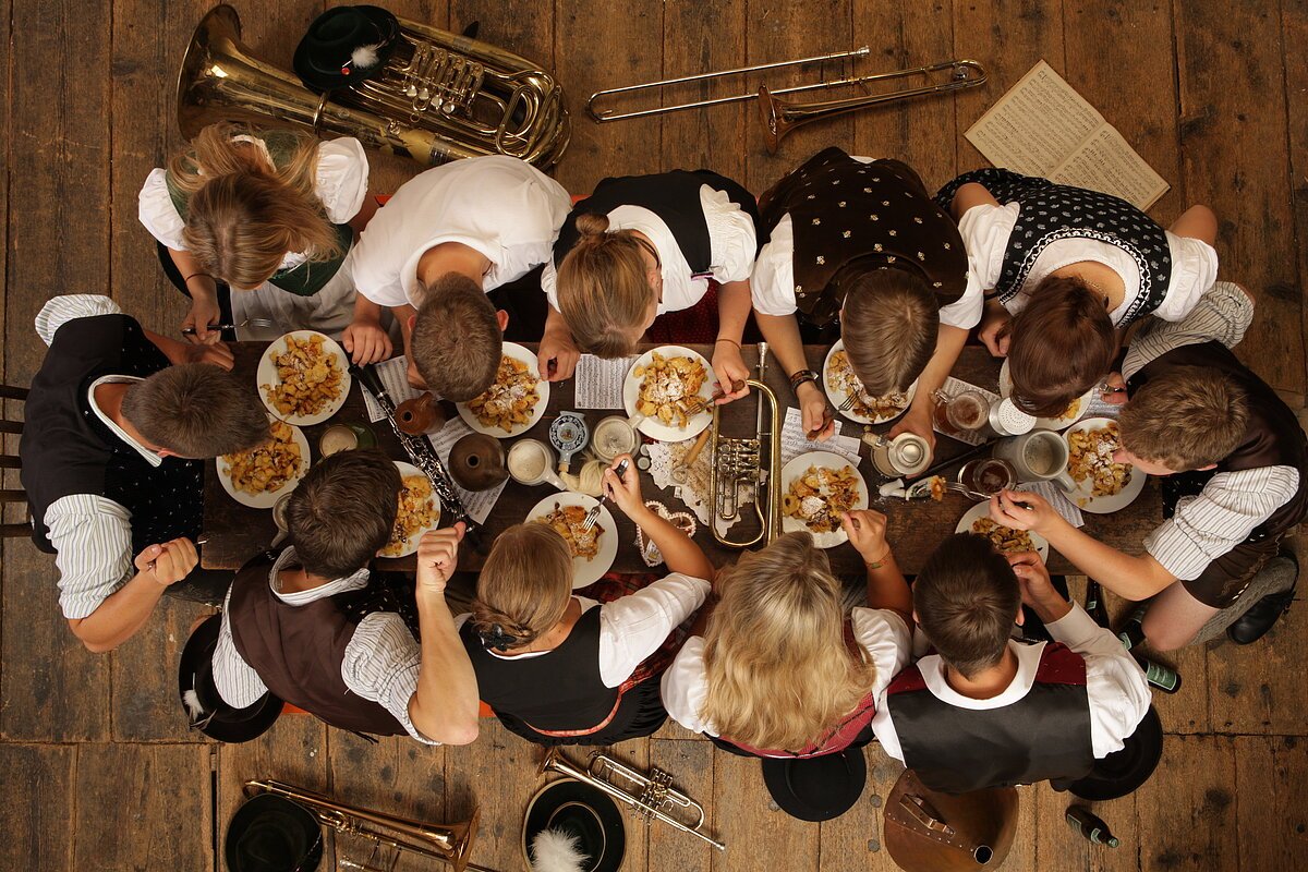 Kapelle Kaiserschmarrn / Fotograf: Hubert Lankes Musikerinnen und Musiker essen in Tracht Kaiserschmarrn an einem Holztisch, umgeben von Instrumenten und Noten.